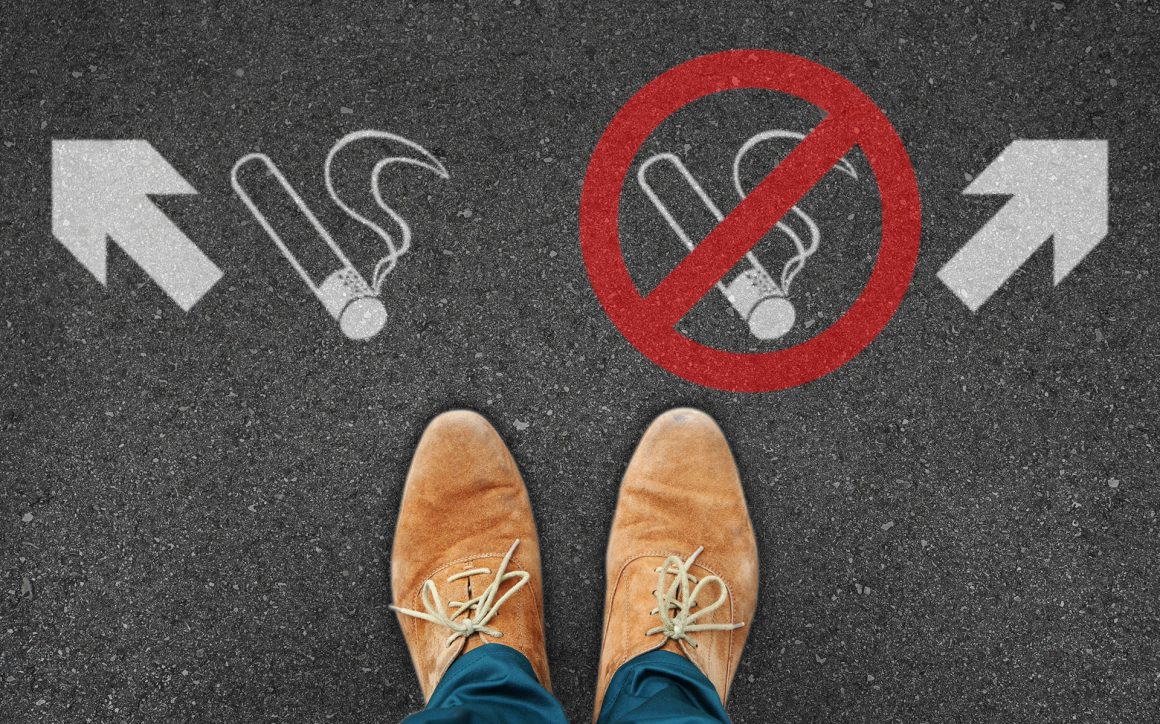Füße, die vor zwei Zeichen stehen, rechts das NIchtraucherzeichen mit Pfeil nach rechts, links ein Raucherzeichen mit Pfeil nach links