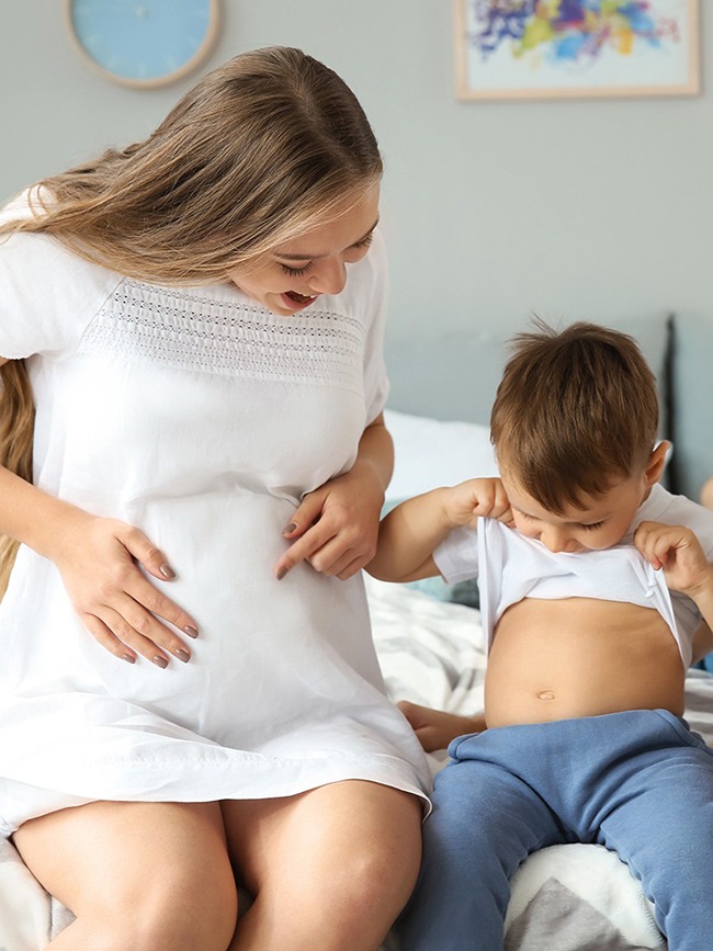 Junge schwangere Frau mit ihrer Familie im Schlafzimmer und das Kleinkind schaut sich seinen eigenen Bauch an, der nicht kugelrund durch eine Schwangerschaft ist.